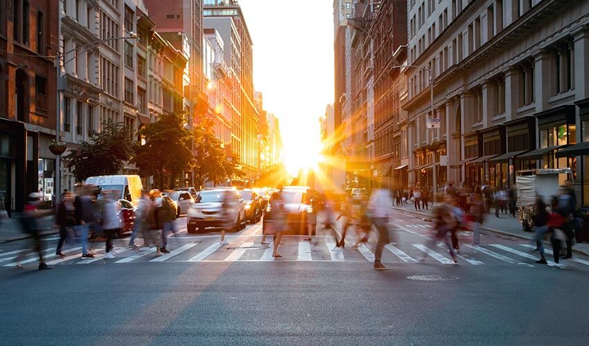 Pedestrians crossing a busy street with sunset in the background.